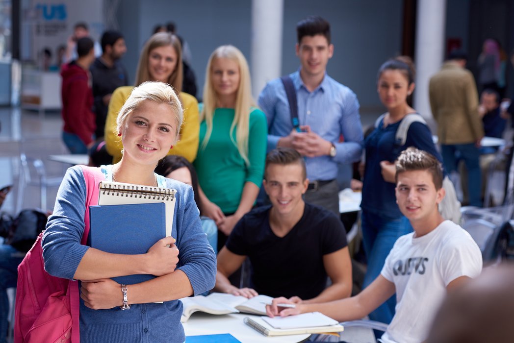Eine junge Frau mit Büchern im Arm lächelt in die Kamera. Im Hintergrund sind weitere Erwachsene, die zusammen an einem Tisch sitzen und lernen. 