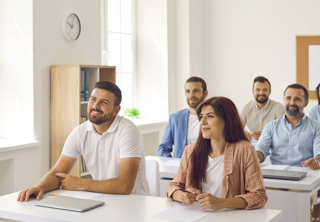 Eine Szene aus der Erwachsenen-Bildung. Eine Lehrerin steht vorn. An Tischen sitzen sechs Erwachsene und hören zu. 