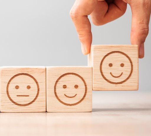In einer Reihe liegen fünf Holzwürfel mit Smileys, die ihren Gesichtsausdruck von Würfel zu Würfel von traurig zu fröhlich ändern. Der Würfel mit dem fröhlichen Gesicht wird von einer Hand angehoben.