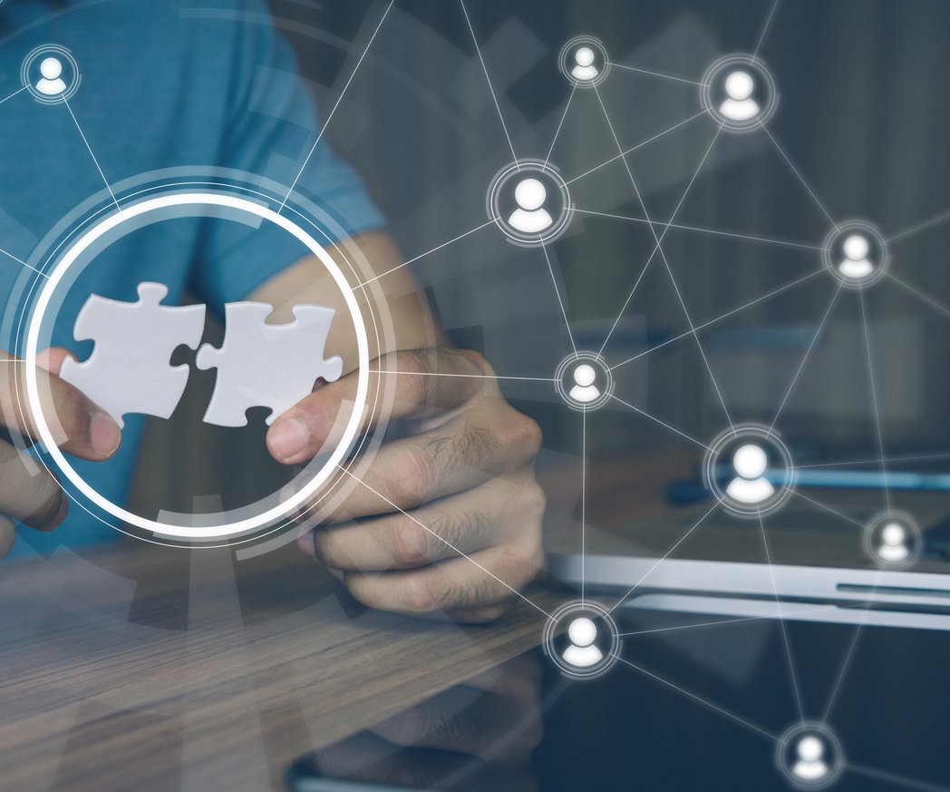 Ein Mann sitz an einem Schreibtisch, vor ihm steht ein Laptop. In seinen Händen hält er zwei grafisch dargestellte Puzzlestücke, die in einander passen. Drum herum sind Grafiken von Personen, die mit einander vernetzt sind.