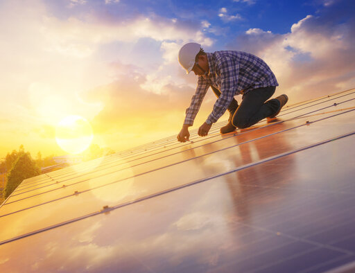 Ein Techniker hockt auf einem Dach und schließt Solar-Paneele an. Im Hintergrund geht die Sonne unter. 
